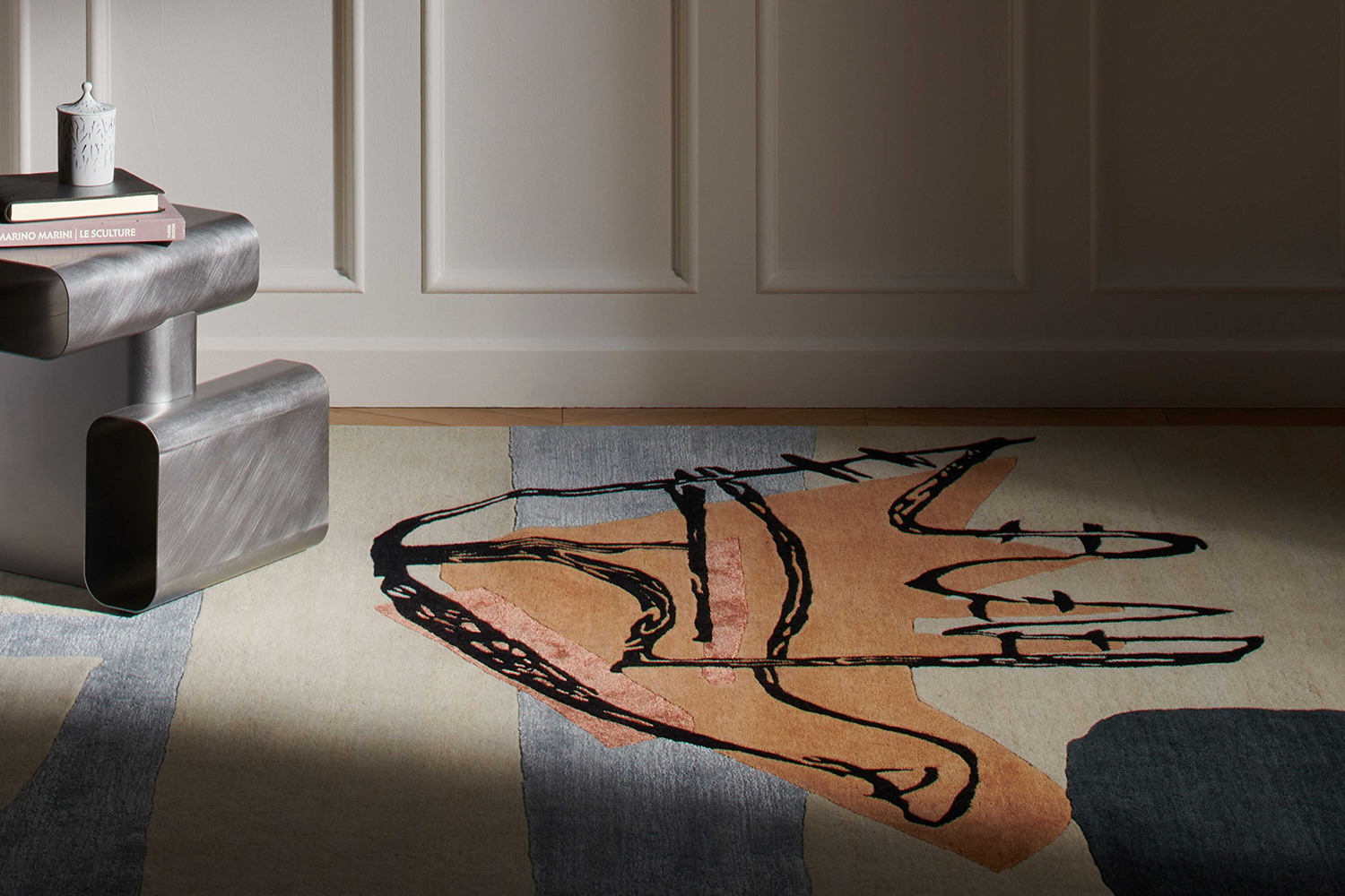 Der Teppich Tapis La Main Ouverte und der Beistelltisch Soft Corners vor einer Stuckwand aus Holz in Weiss. 